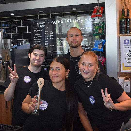 Student staff all posing and smiling at the camera in front of the coffee menu